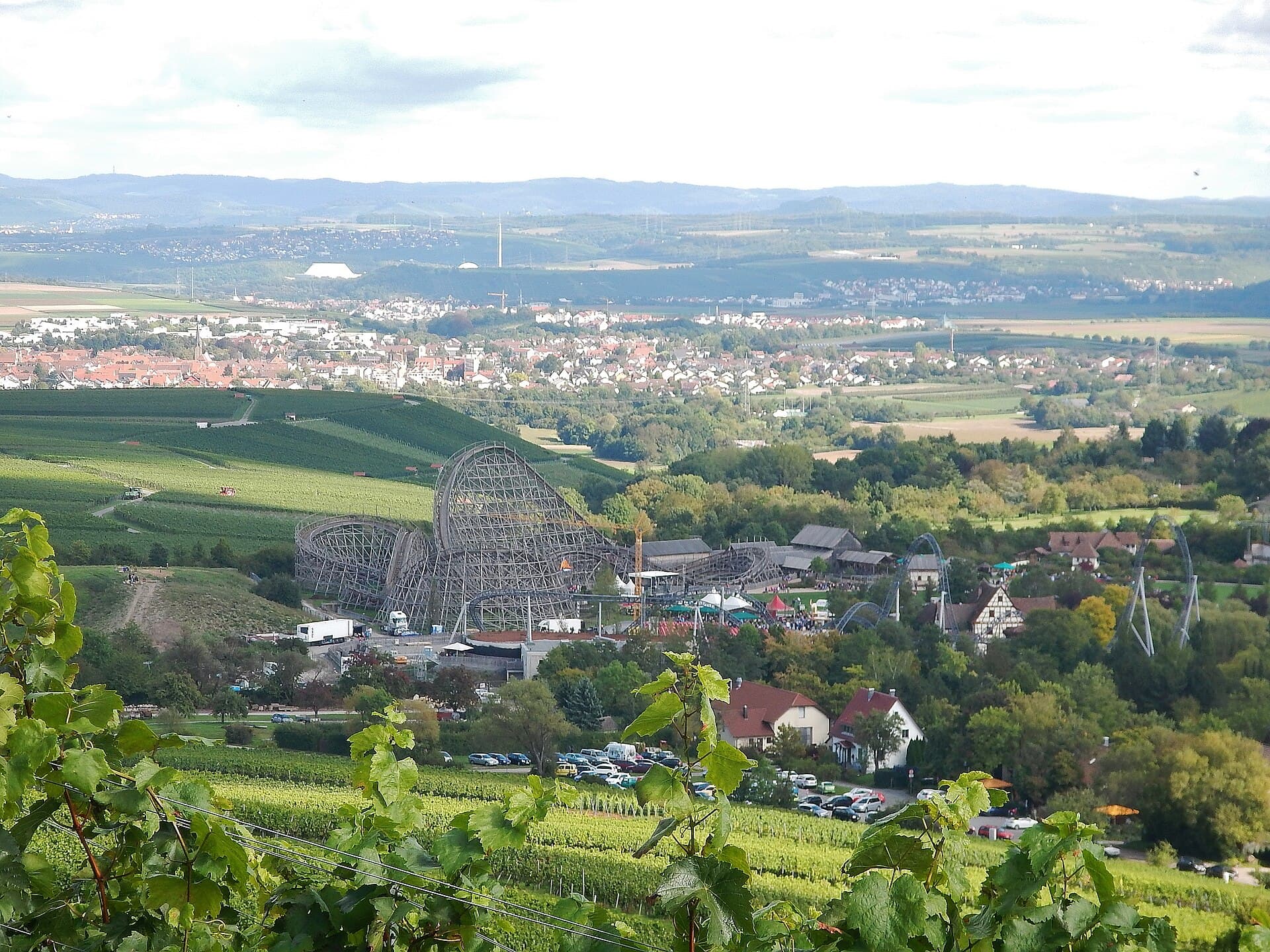 Blick aus den Weinbergen am Michaelsberg auf den Erlebnispark Tripsdrill mit der Holzachterbahn Mammut, dahinter Orte und Felder in Richtung Neckar.