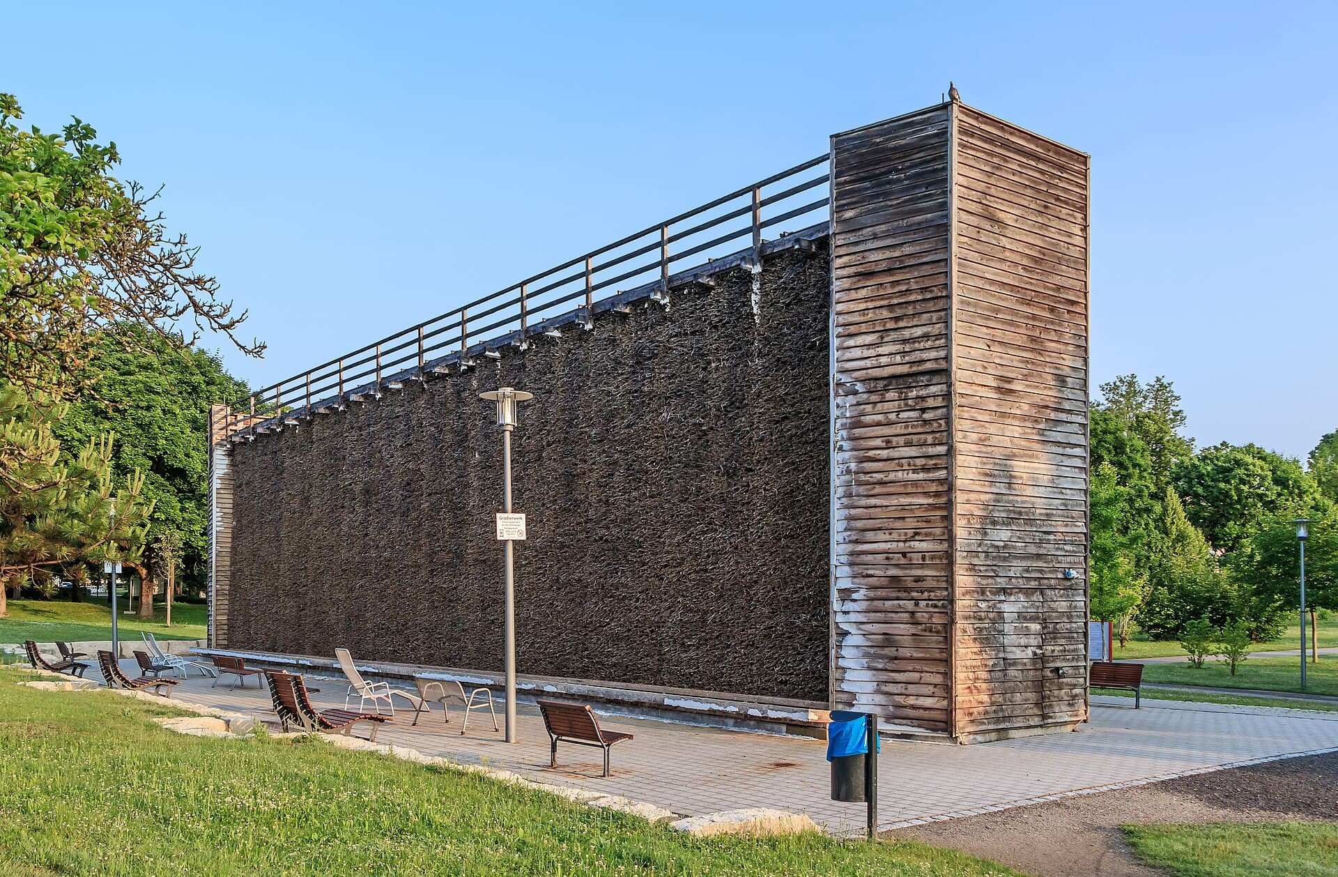 Das Gradierwerk im Salinenpark Bad Rappenau: eine lange Wand aus dunklem Schlehenreisig mit hölzernem Eckturm und Geländer, davor ein gepflasterter Platz mit Bänken und Liegestühlen.