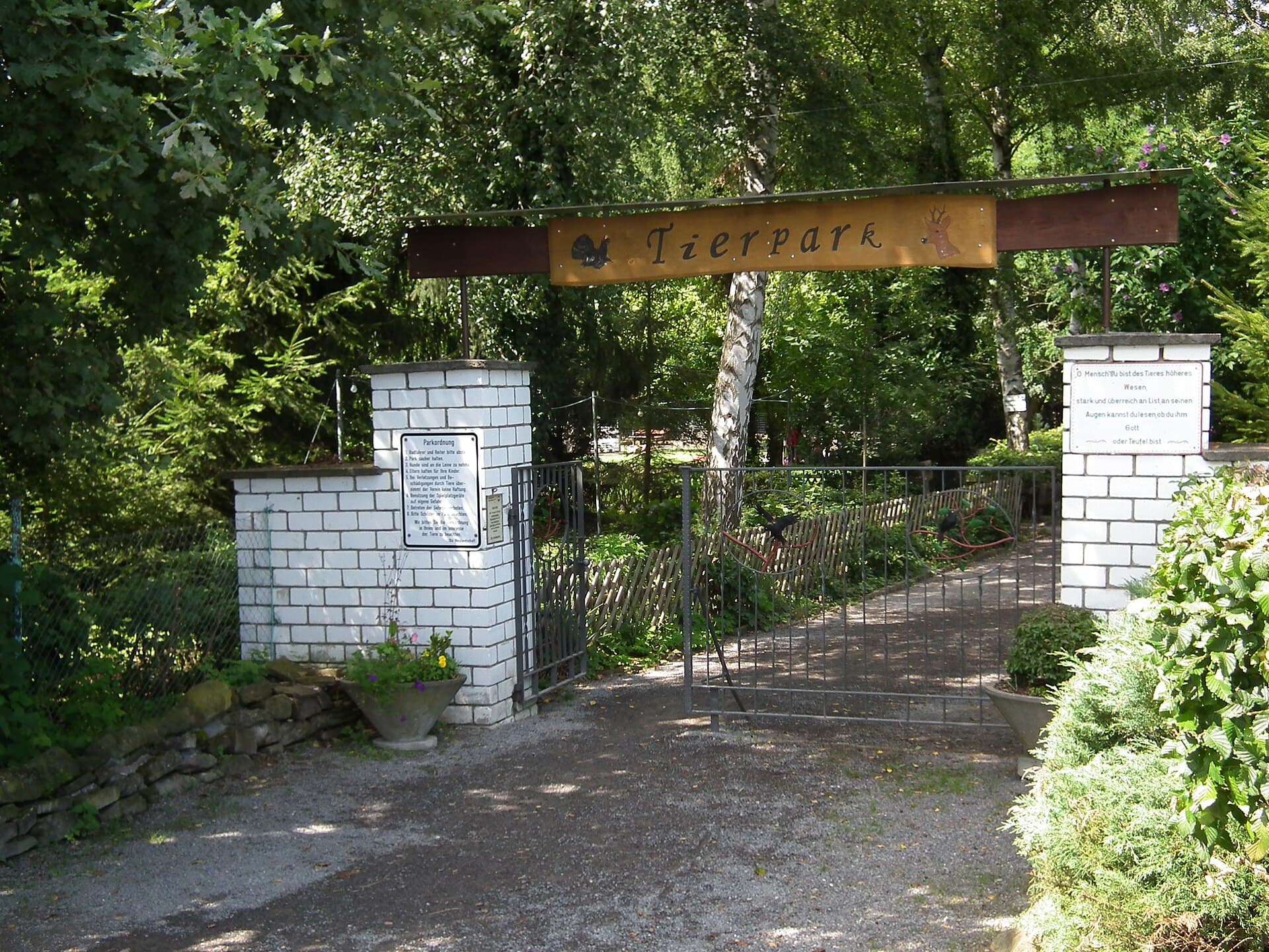 Der Haupteingang des Kleintierparks: ein offenes Gittertor zwischen weiß gemauerten Pfeilern unter einem Holzschild mit der Aufschrift „Tierpark“, davor ein Kiesvorplatz, dahinter Bäume und ein Weg in den Park.