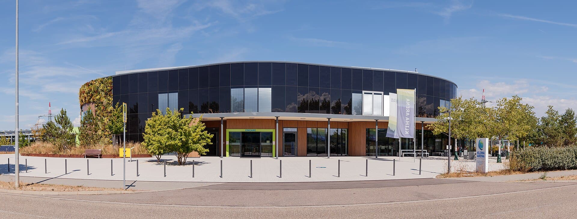 Das runde Gebäude der KLIMA ARENA von der Dietmar-Hopp-Straße aus, mit dunkler Fassade, begrünter Wand am linken Rand und dem Haupteingang hinter dem Vorplatz