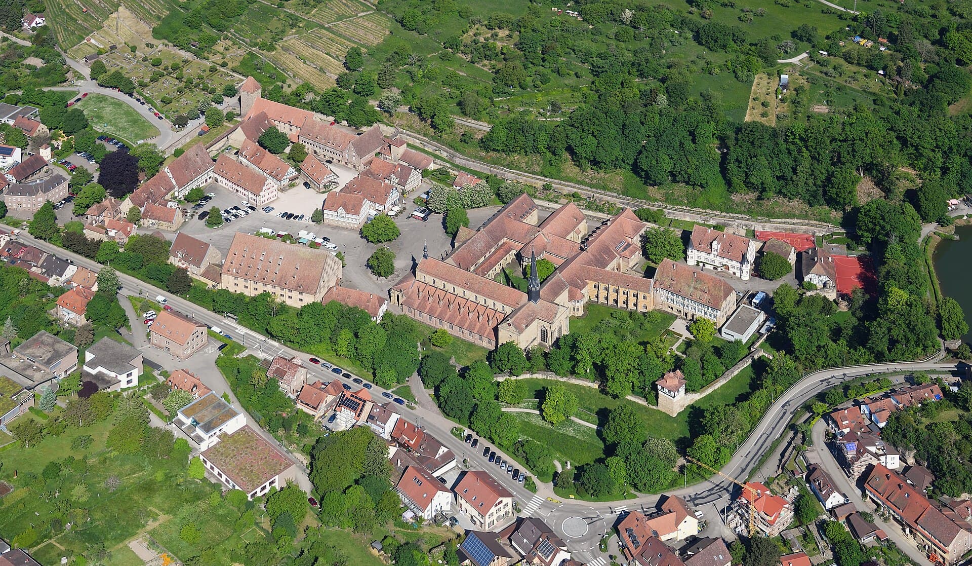 Luftbild von Südosten auf Kloster Maulbronn mit der Klosterkirche und ihrem spitzen Dachreiter in der Mitte, links dem Klosterhof mit Fruchtkasten und Fachwerkhäusern, vorn der Klostermauer mit einem Turm, oben Weinbergen am Hang und am rechten Rand dem Tiefen See.