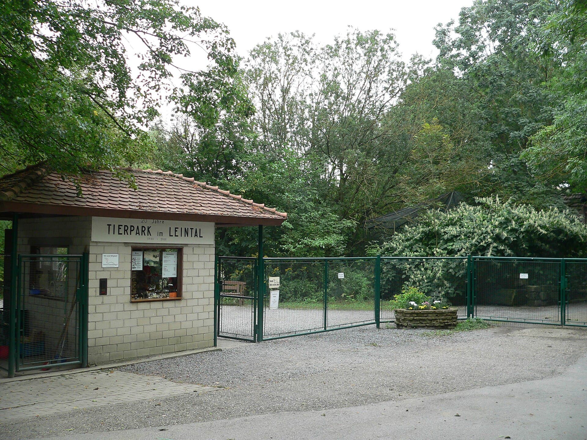 Das Kassenhaus mit der Aufschrift „Tierpark im Leintal“ und daneben das grüne Eingangstor des Leintalzoos, dahinter hohe Bäume, im Vordergrund der geschotterte Vorplatz.