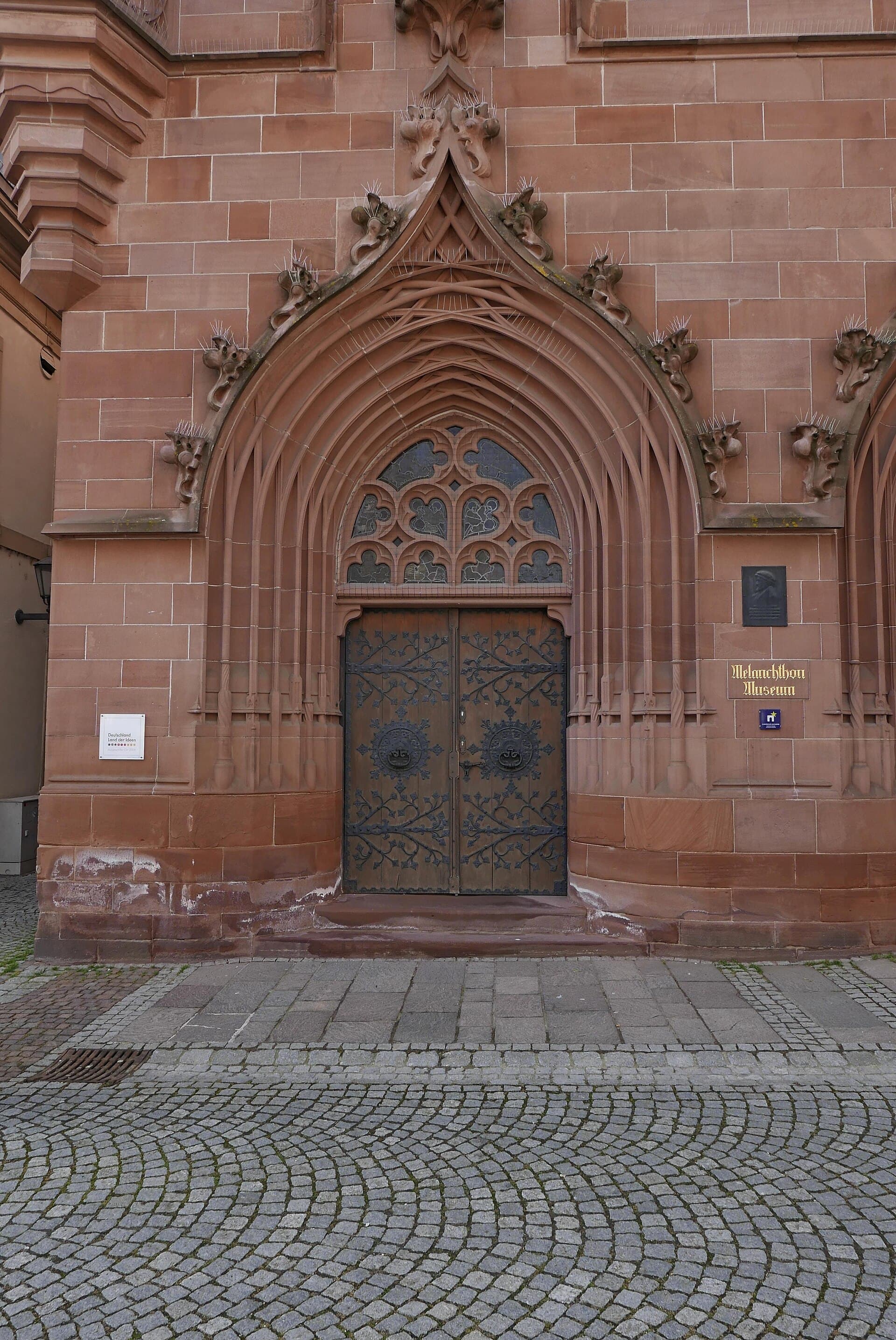 Spätgotisch gestaltetes Portal aus rotem Sandstein am Melanchthonhaus in Bretten mit schmiedeeisern beschlagener Holztür und dem Schild „Melanchthon Museum“, davor das Pflaster des Marktplatzes.