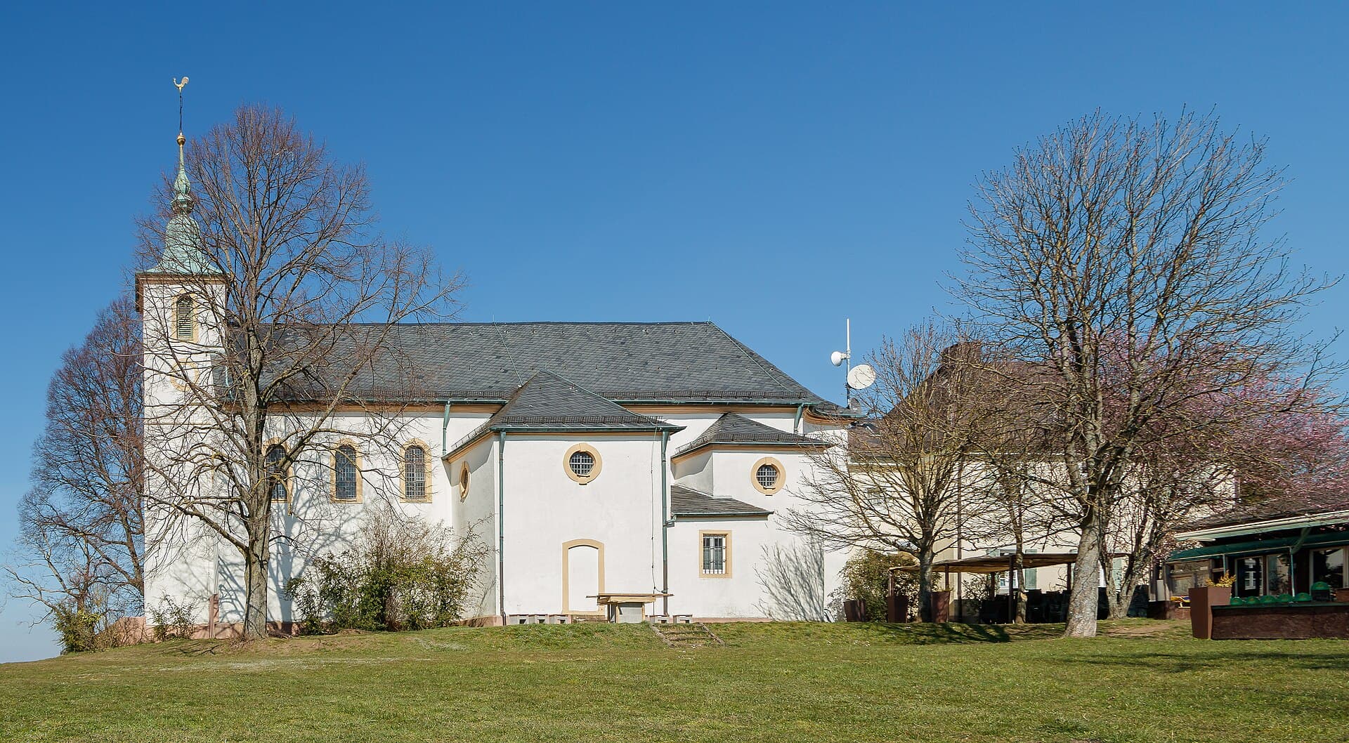 Die Michaelskapelle auf dem Michaelsberg von Süden: weiß verputzter Barockbau mit Turm und grüner Haube hinter einer Wiese mit kahlen Bäumen, rechts das Nebengebäude mit der Terrasse des Restaurants.