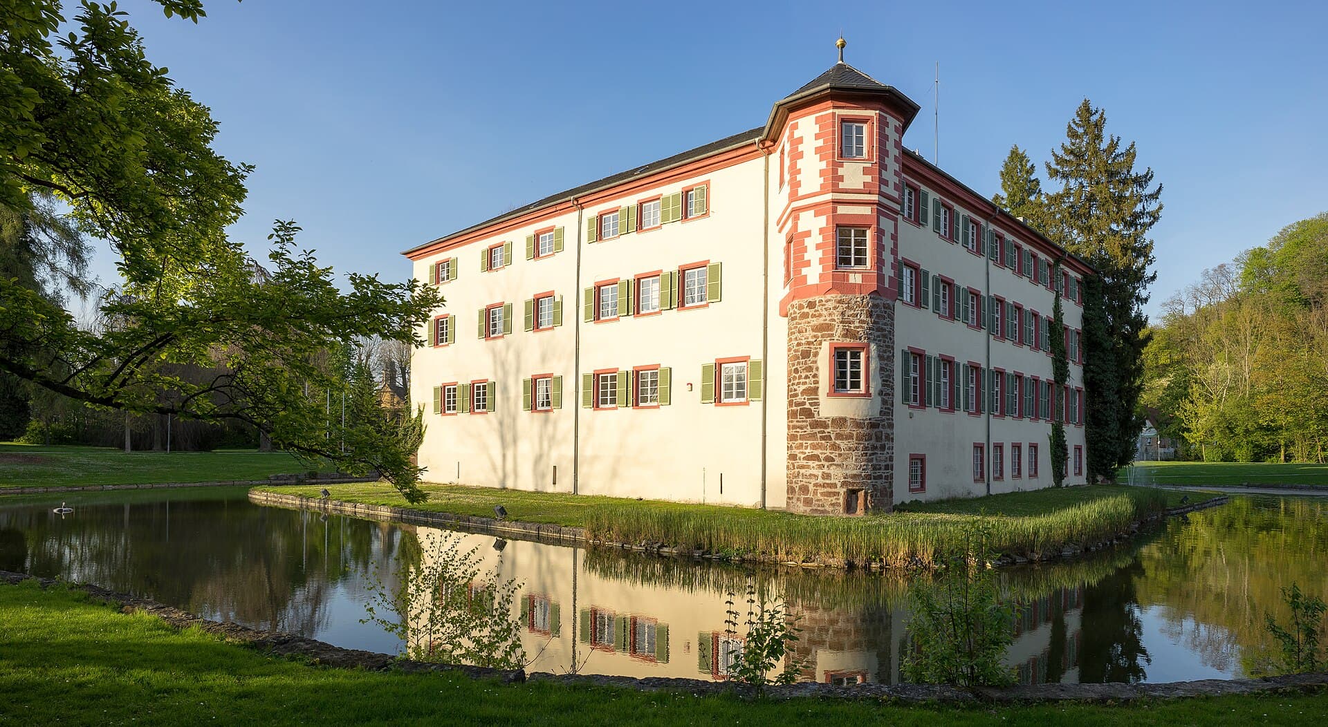 Schloss Eichtersheim von Südwesten: der runde Eckturm und die hellen Fassaden spiegeln sich im Schlossteich, davor Rasen und Bäume des Schlossparks.
