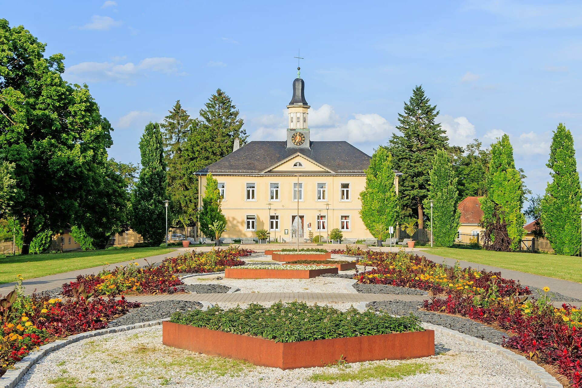 Das gelbe Salinenamtsgebäude von 1823 mit Uhrtürmchen im Salinenpark von Bad Rappenau, davor das Feuerbeet mit roten und gelben Sommerblumen