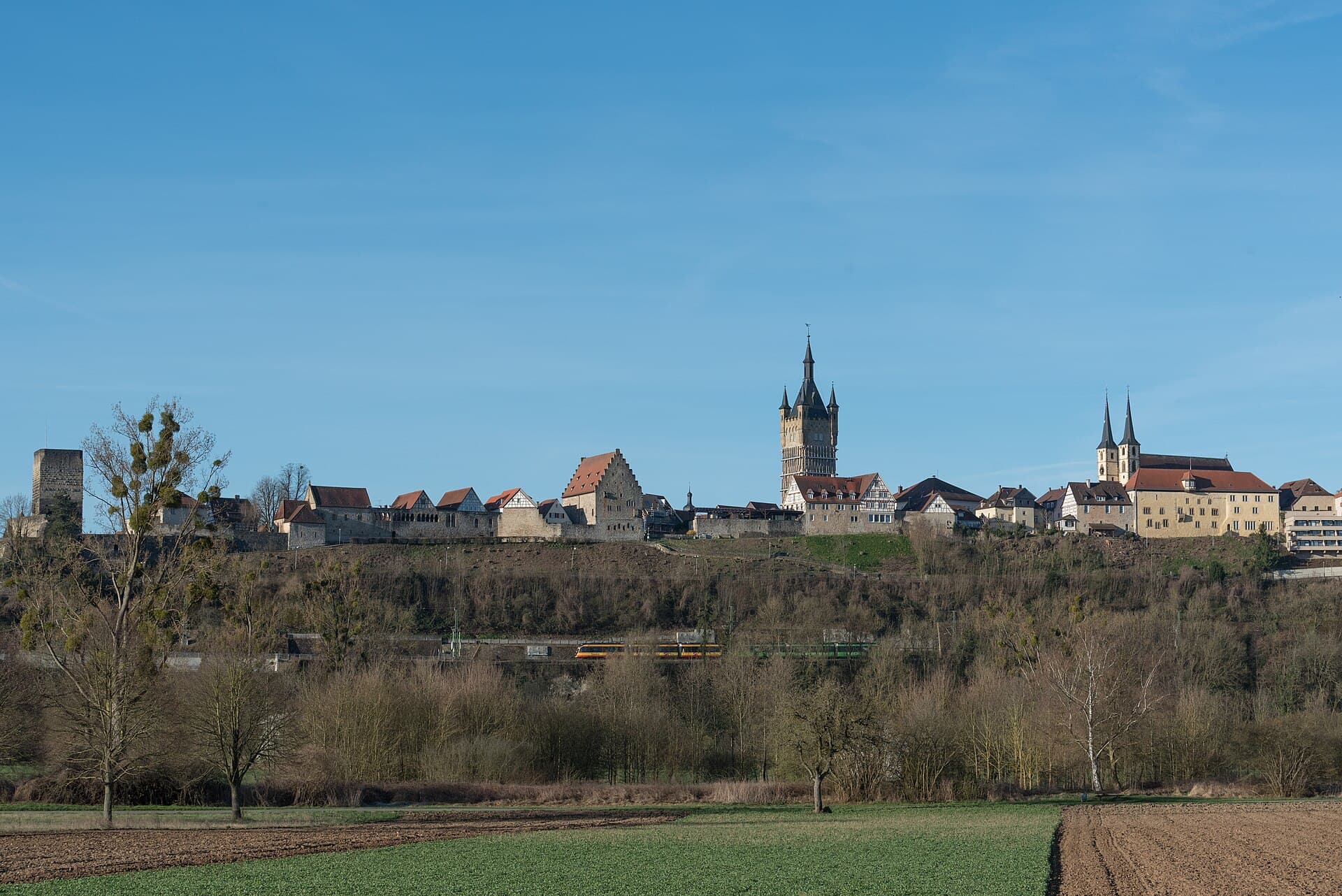 Die Altstadt von Bad Wimpfen am Berg von Nordosten mit Rotem Turm, Blauem Turm und den Türmen der Stadtkirche über dem Hang, darunter Bahnstrecke und Felder