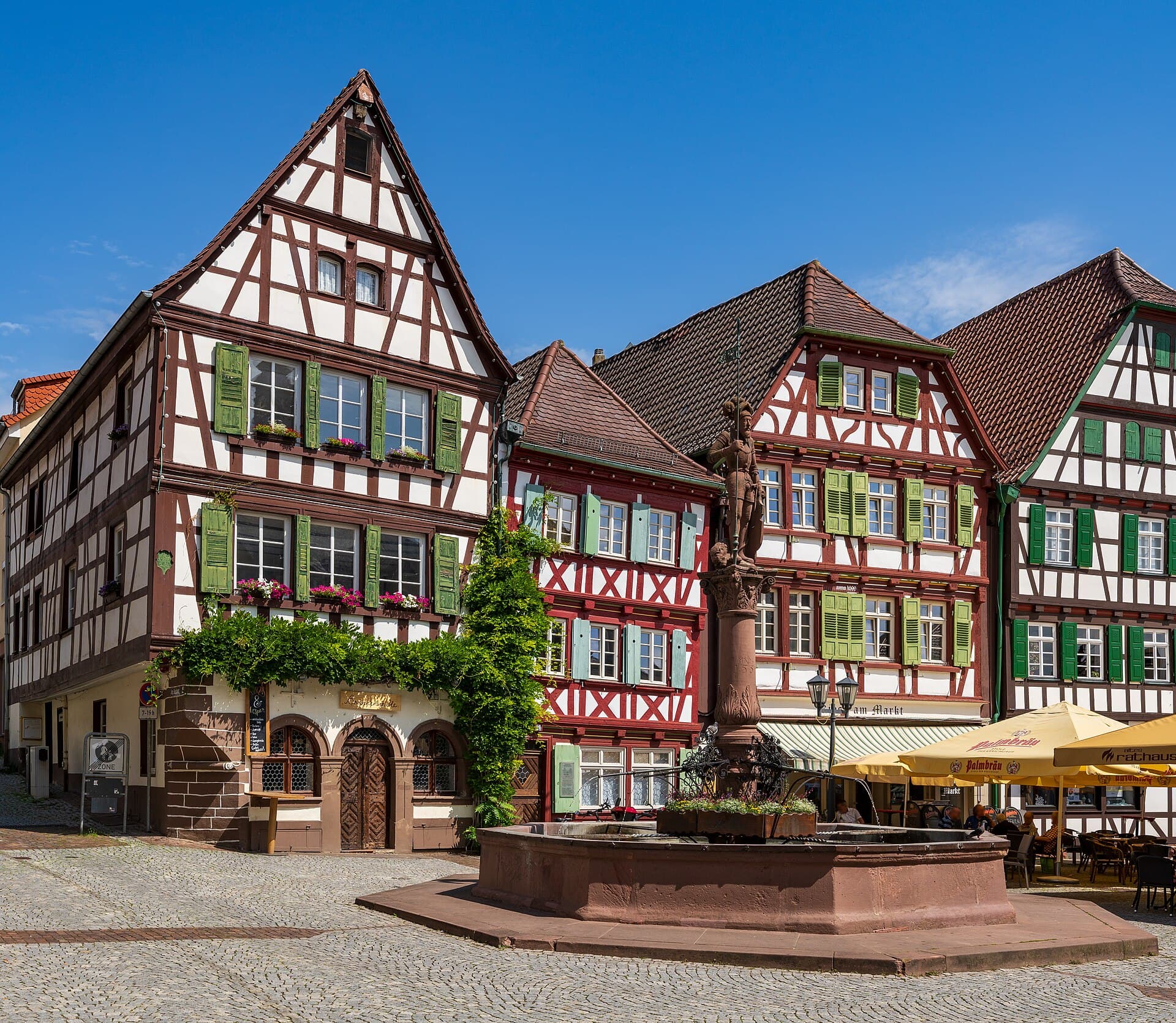 Fachwerkhäuser mit grünen Fensterläden am Marktplatz von Bretten, davor der Marktbrunnen mit der Säulenfigur des Kurfürsten Friedrich II. von der Pfalz unter blauem Himmel