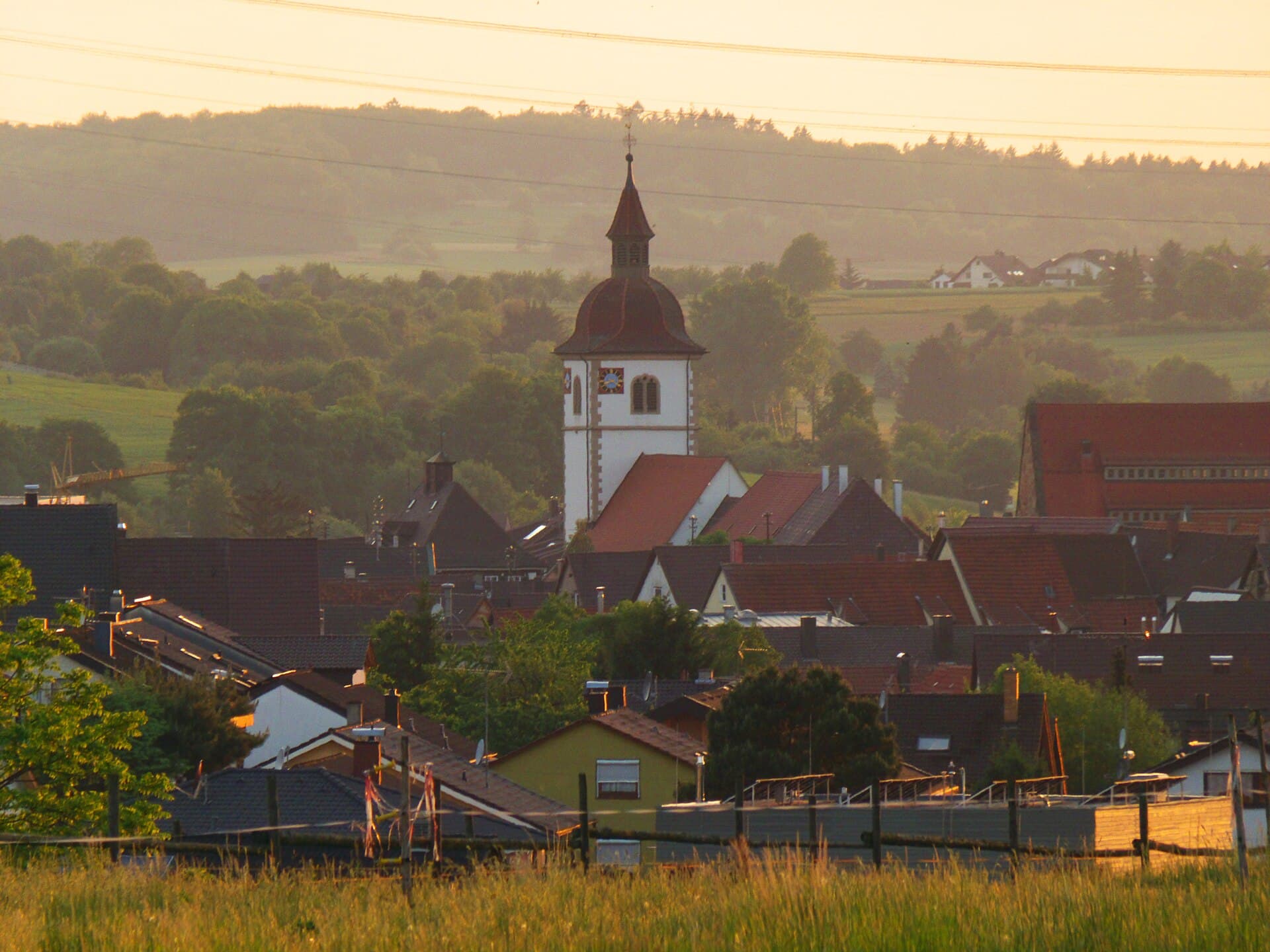 Blick über die Dächer von Knittlingen auf den weißen Turm der St. Leonhardskirche mit Uhr und rotbrauner Haube, dahinter Wiesen und bewaldete Hügel im Abendlicht