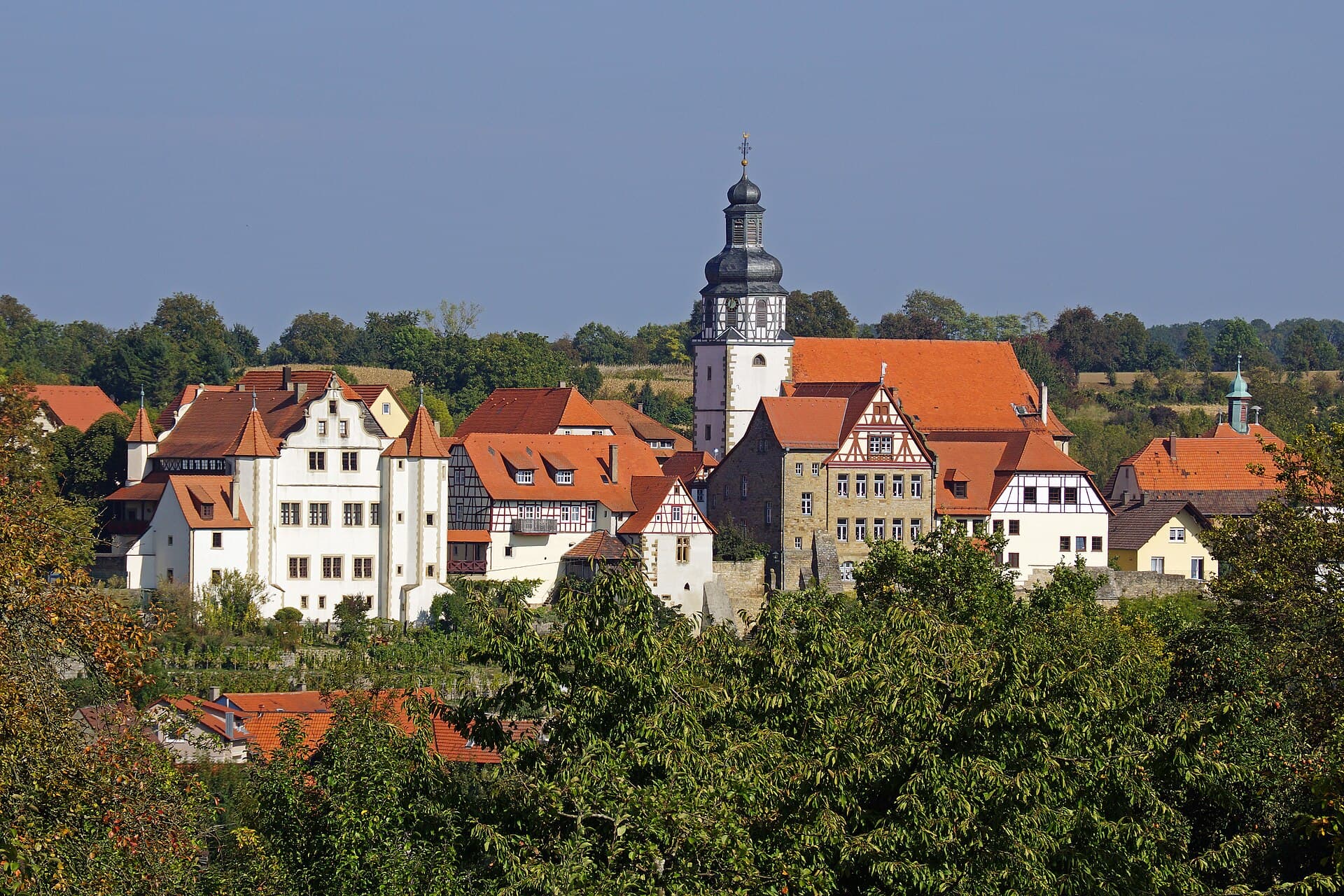 Blick über Baumkronen auf die Altstadt von Gochsheim mit dem weißen Graf-Eberstein-Schloss, dem Turm der Martinskirche mit dunkler Haube und Fachwerkhäusern unter roten Dächern