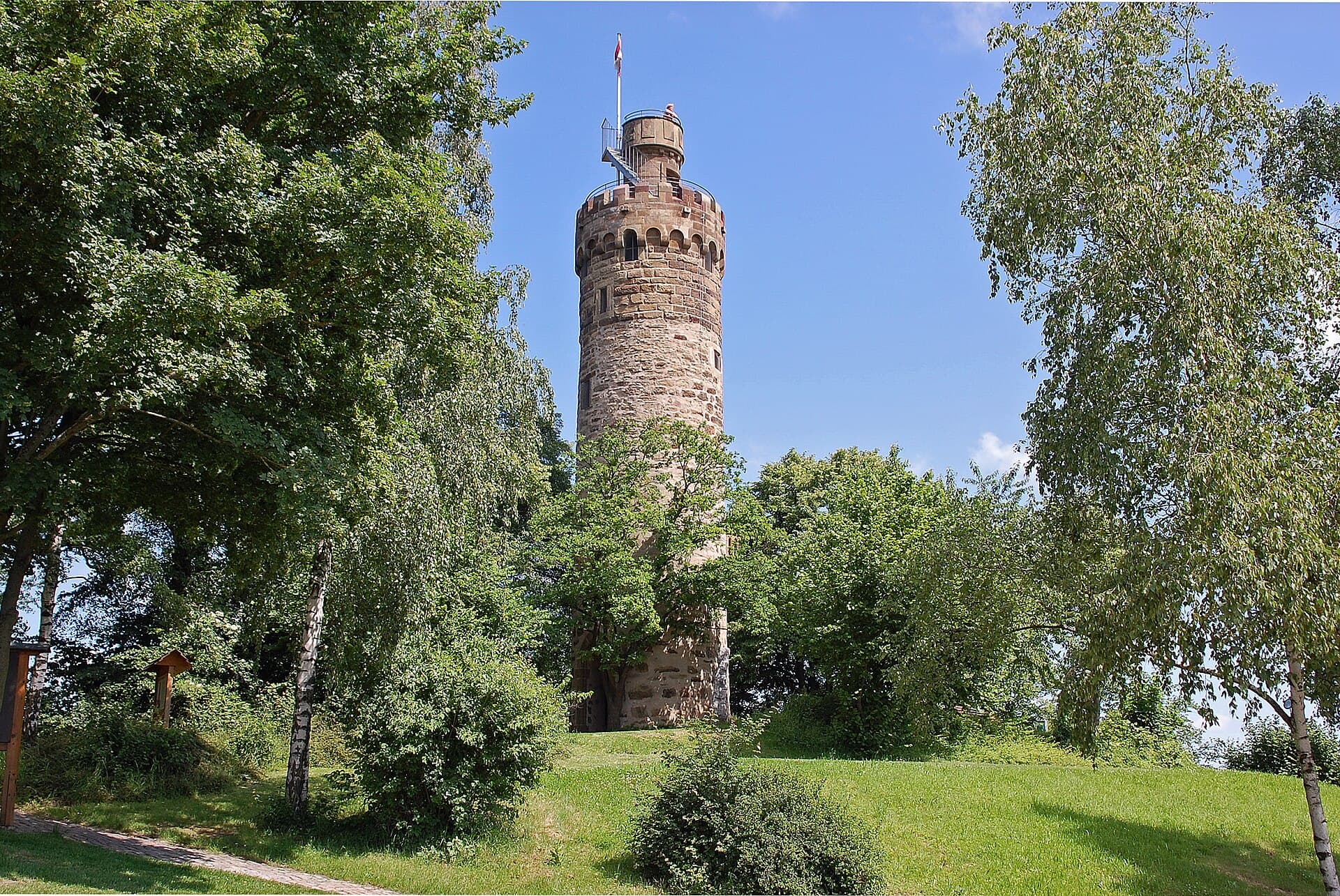 Der runde Sandsteinturm der Heuchelberger Warte mit Zinnenkranz und Fahne ragt hinter Bäumen über eine gemähte Wiese in den blauen Himmel