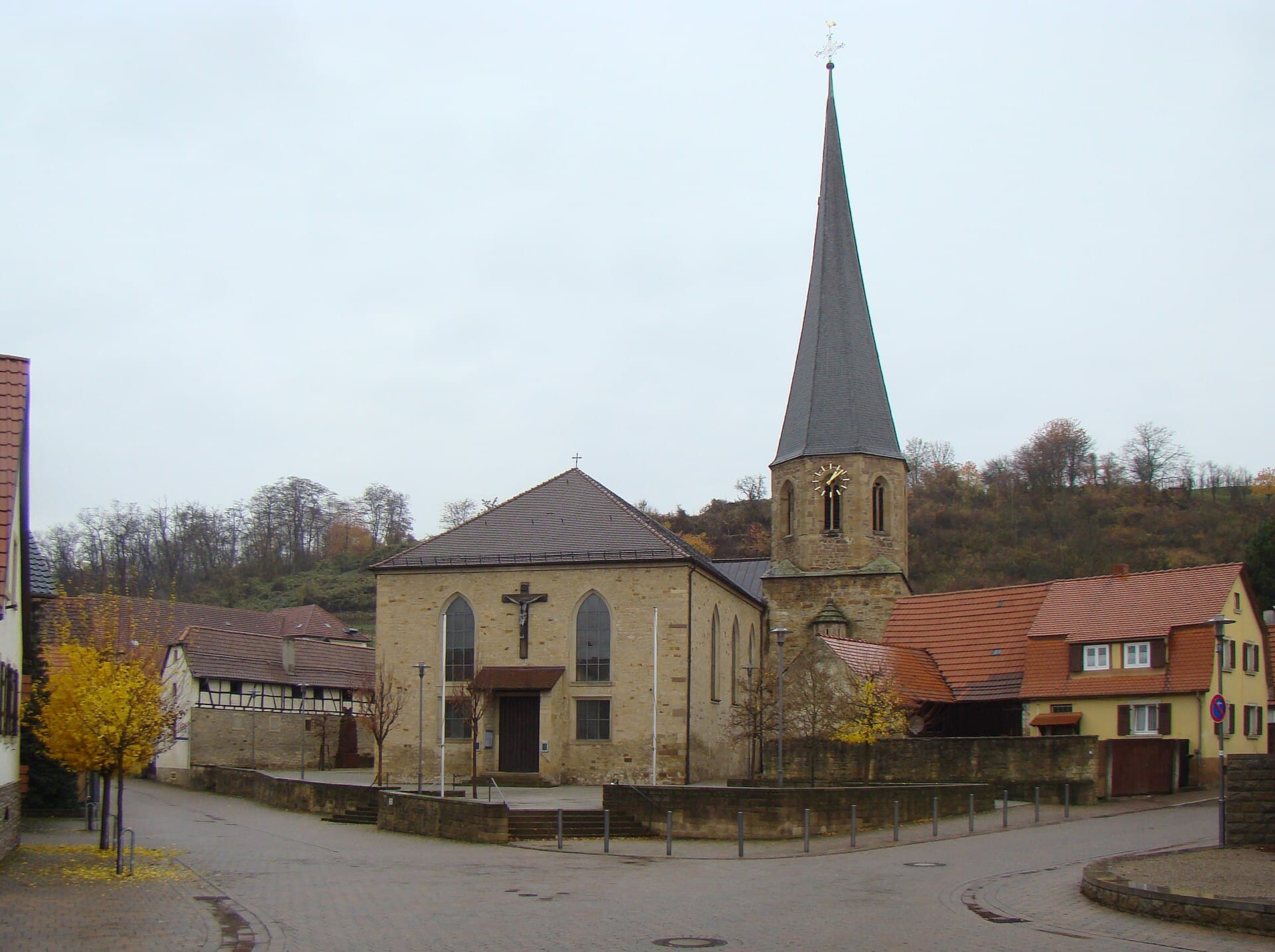 Die katholische Kirche St. Cäcilia in Mühlhausen mit spitzem Turmhelm über dem gepflasterten Kirchplatz, dahinter ein Hang mit herbstlichen Bäumen