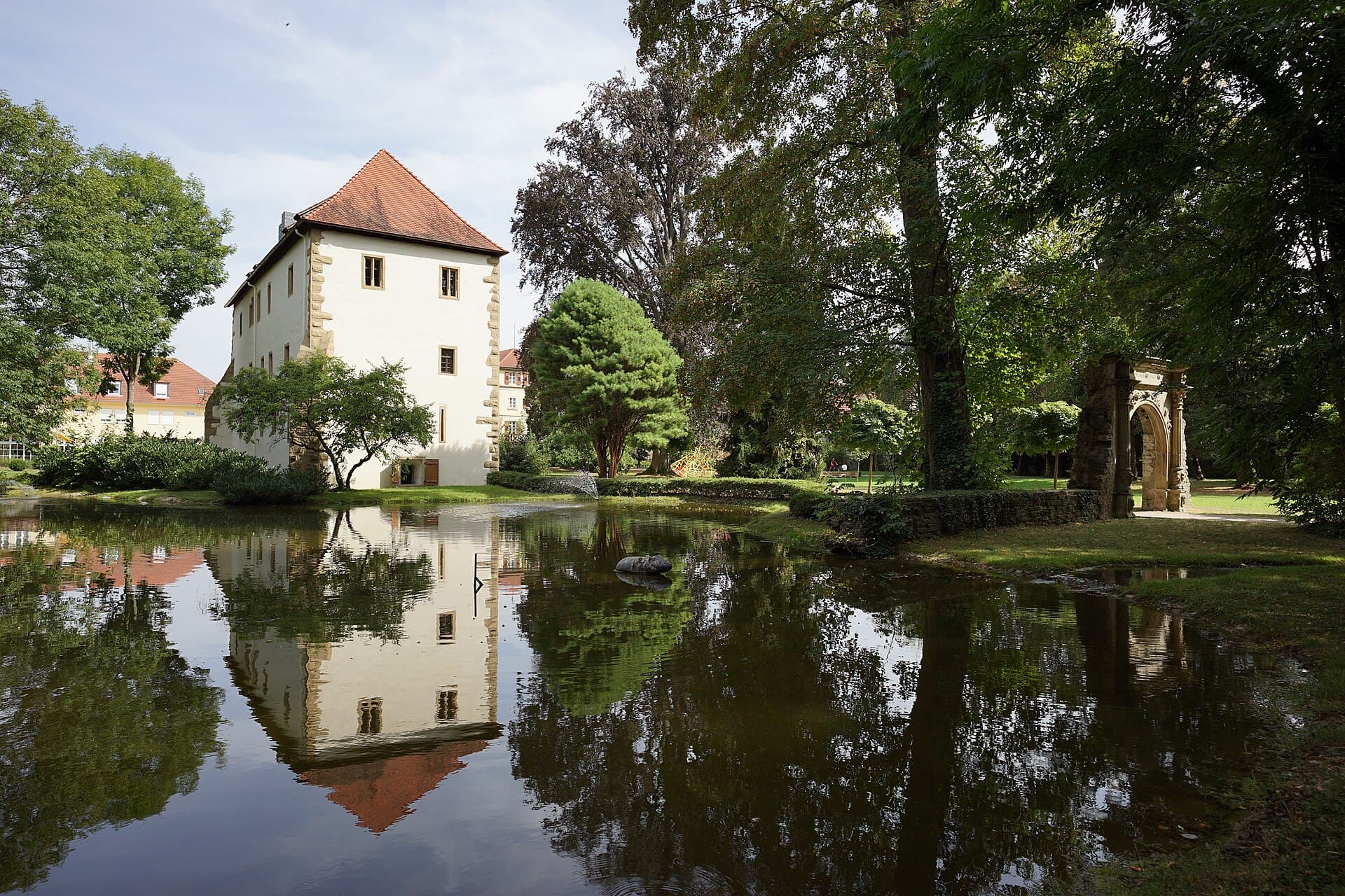 Das weiß verputzte Alte Schloss mit rotem Walmdach spiegelt sich im Schlossteich, rechts steht unter hohen Bäumen ein steinernes Renaissance-Tor im Schlosspark.
