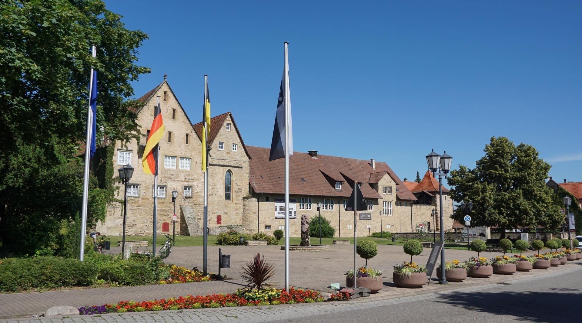 Blick über den Marktplatz von Oberderdingen auf die Sandsteinbauten des Amthofs mit hohem Giebelhaus, Spitzbogenfenster und einem Eckturm mit rotem Dach, davor Fahnenmasten, Laternen und Blumenkübel