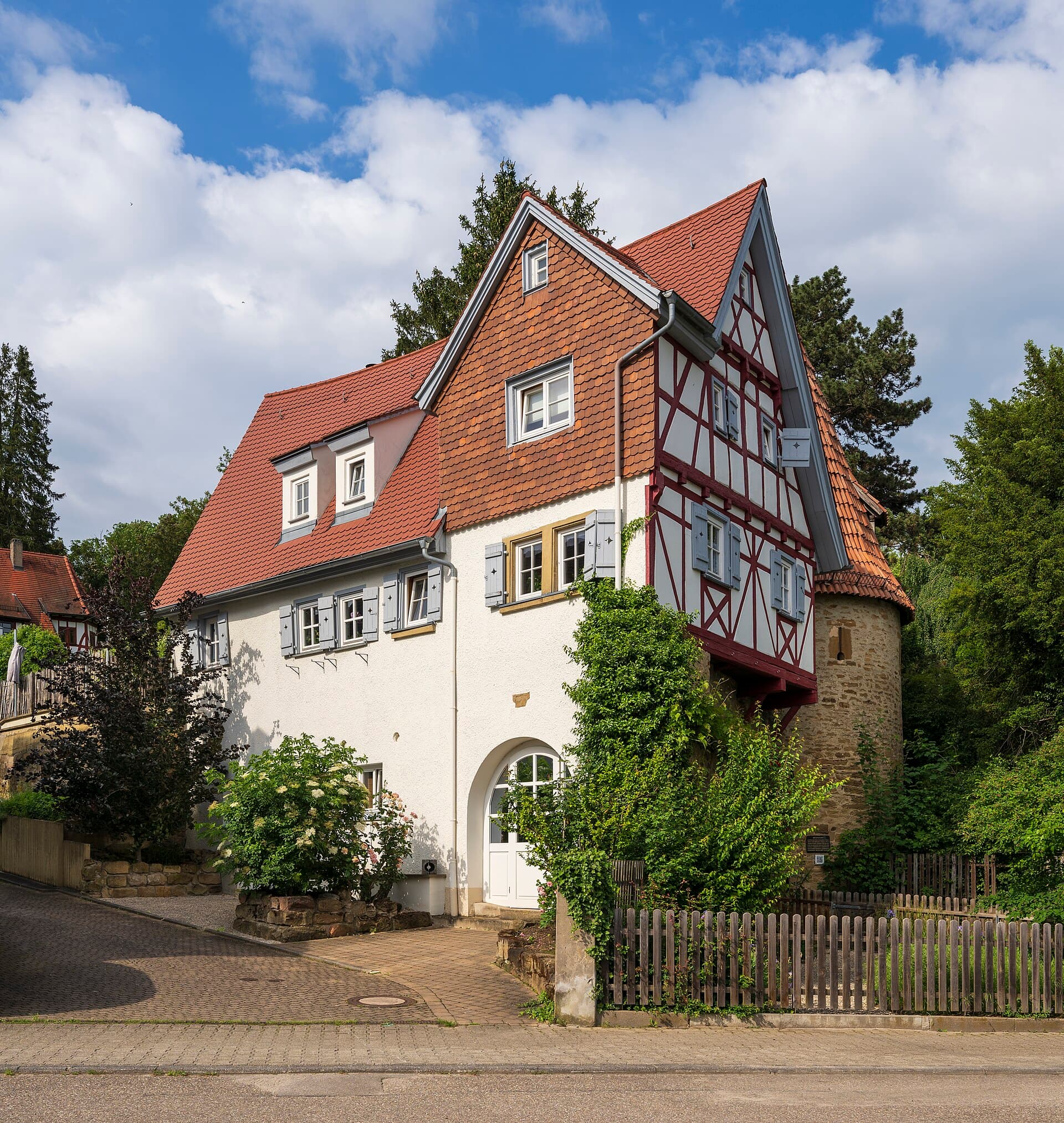 Das weiß verputzte Reinwaldsche Haus mit rotbraun verschindeltem Giebel und Fachwerk am Hindenburgplatz in Schwaigern, rechts angebaut der runde Hexenturm aus Sandstein, davor ein Holzzaun unter blauem Himmel mit Wolken