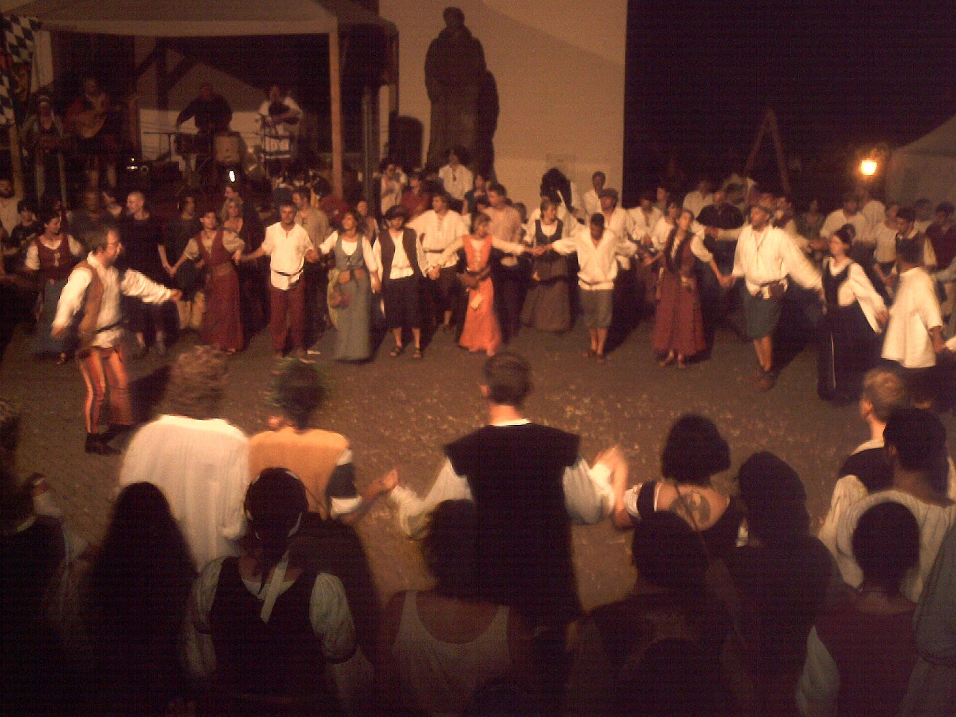Mitwirkende in historischen Gewändern tanzen nachts beim Peter-und-Paul-Fest 2009 einen Reigen auf dem Kirchplatz in Bretten, dahinter spielt eine Musikgruppe auf einer Bühne.