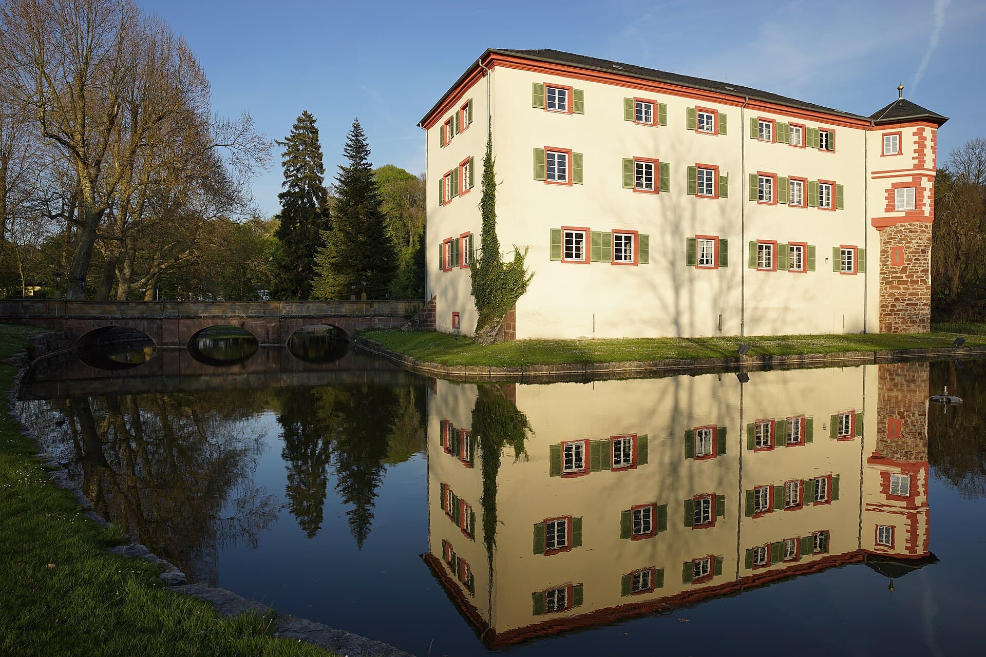 Das Wasserschloss Eichtersheim von Westen an einem sonnigen Frühlingsabend, die Fassade spiegelt sich im Schlossteich, links führt die Sandsteinbrücke über das Wasser.