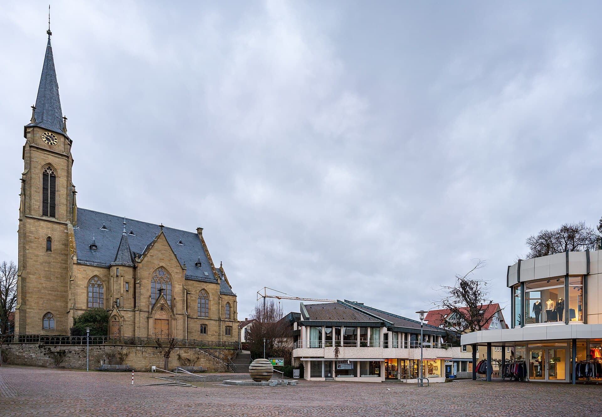 Der Kirchplatz in Bad Rappenau an einem trüben Januarabend: links die evangelische Stadtkirche, in der Mitte ein Brunnen mit einer Kugel aus Steinscheiben vor dem evangelischen Gemeindehaus, rechts ein beleuchtetes Modegeschäft.
