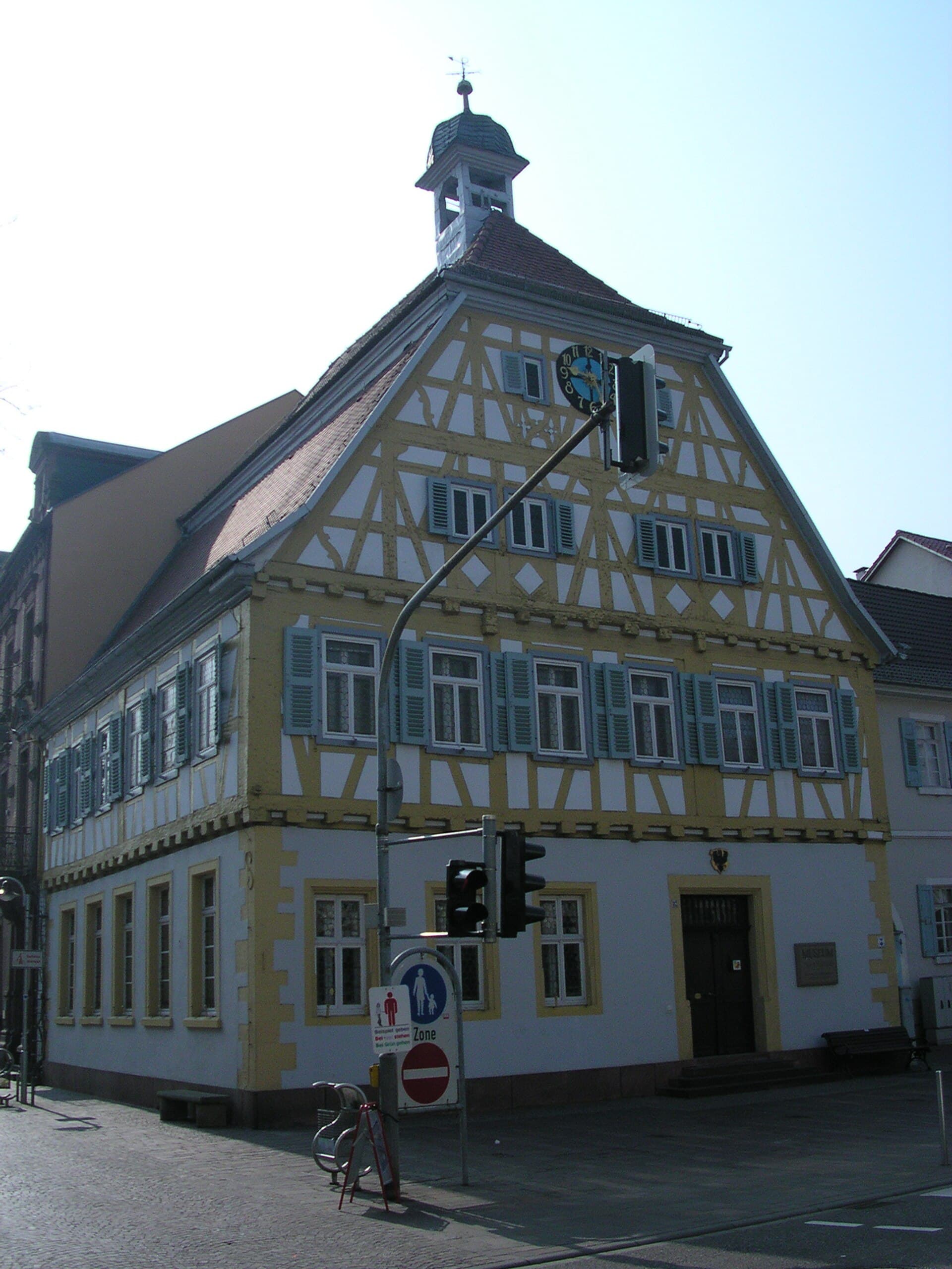 Das Alte Rathaus an der Sinsheimer Hauptstraße, ein Fachwerkbau mit gelb gefassten Balken, blaugrauen Fensterläden, Uhr im Giebel und kleinem Dachreiter, davor die leere Fußgängerzone mit Ampel und Verkehrszeichen.
