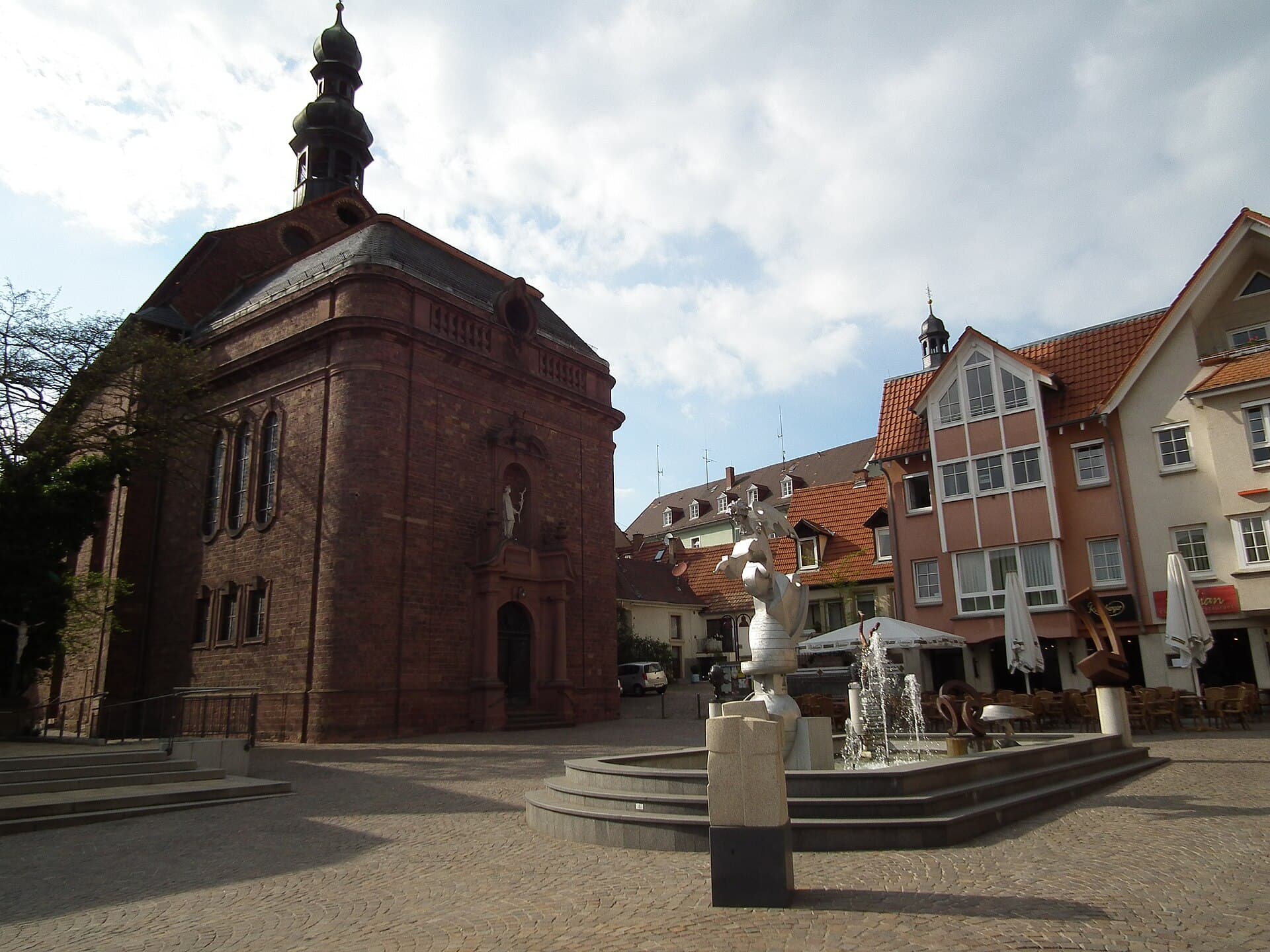 Der Adenauerplatz in Wiesloch mit der Brunnengalerie, links die rote Sandsteinfassade der katholischen Kirche St. Laurentius, rechts Cafétische und Sonnenschirme vor einer Häuserzeile.