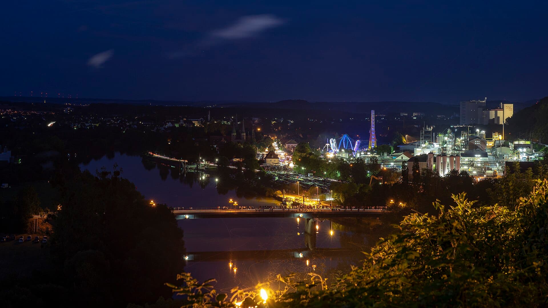 Blick von der Altstadt am Berg auf Wimpfen im Tal am Abend des Talmarkts 2018: vorne Neckar und Brücke, dahinter die Türme der Ritterstiftskirche St. Peter und die beleuchteten Fahrgeschäfte, rechts das Solvay-Werk