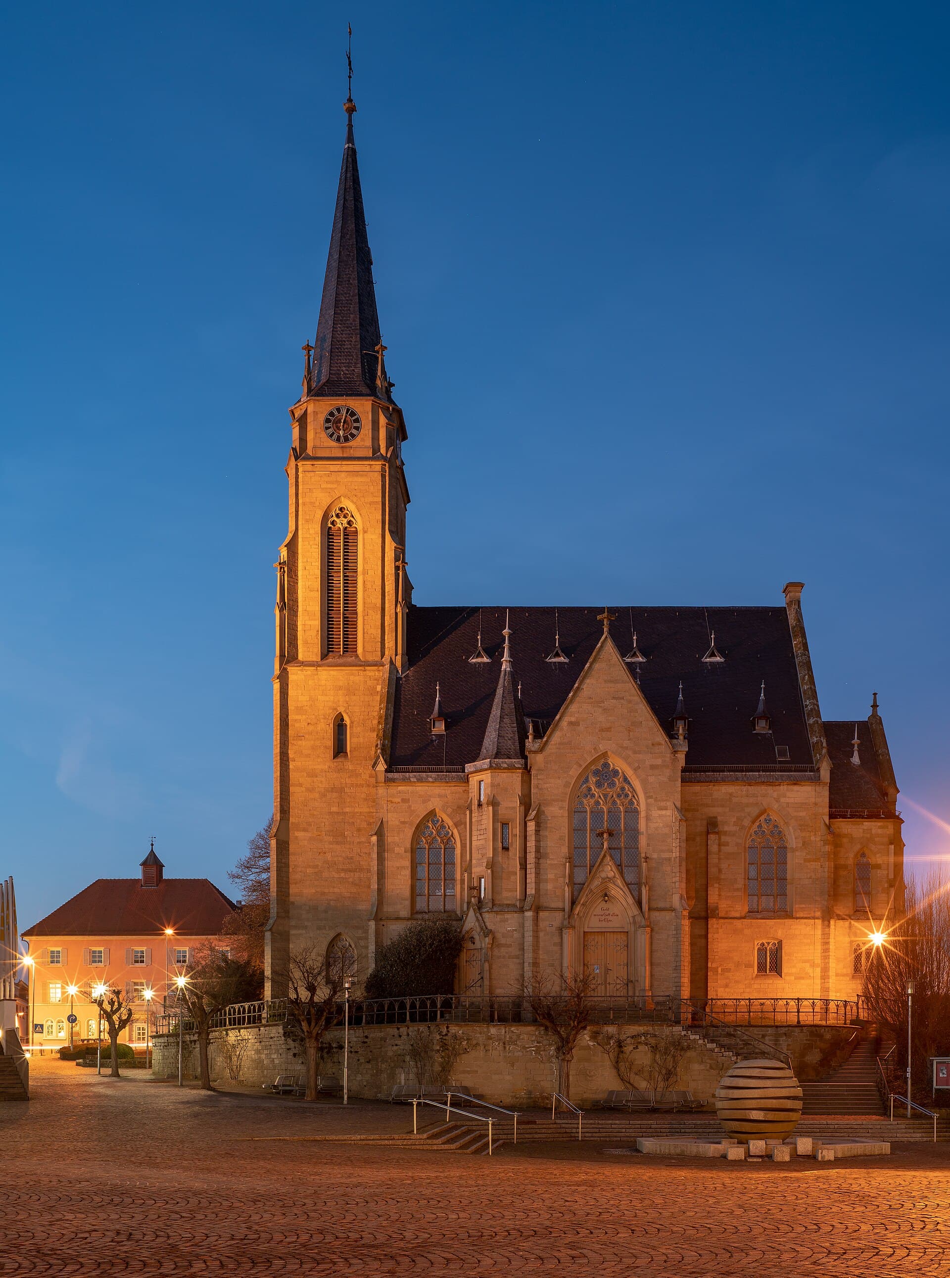 Die angestrahlte evangelische Stadtkirche von Bad Rappenau an einem Winterabend in der blauen Stunde, gesehen vom Kirchplatz, links im Hintergrund das Alte Rathaus und rechts ein Brunnen mit einer Kugel aus Steinscheiben.