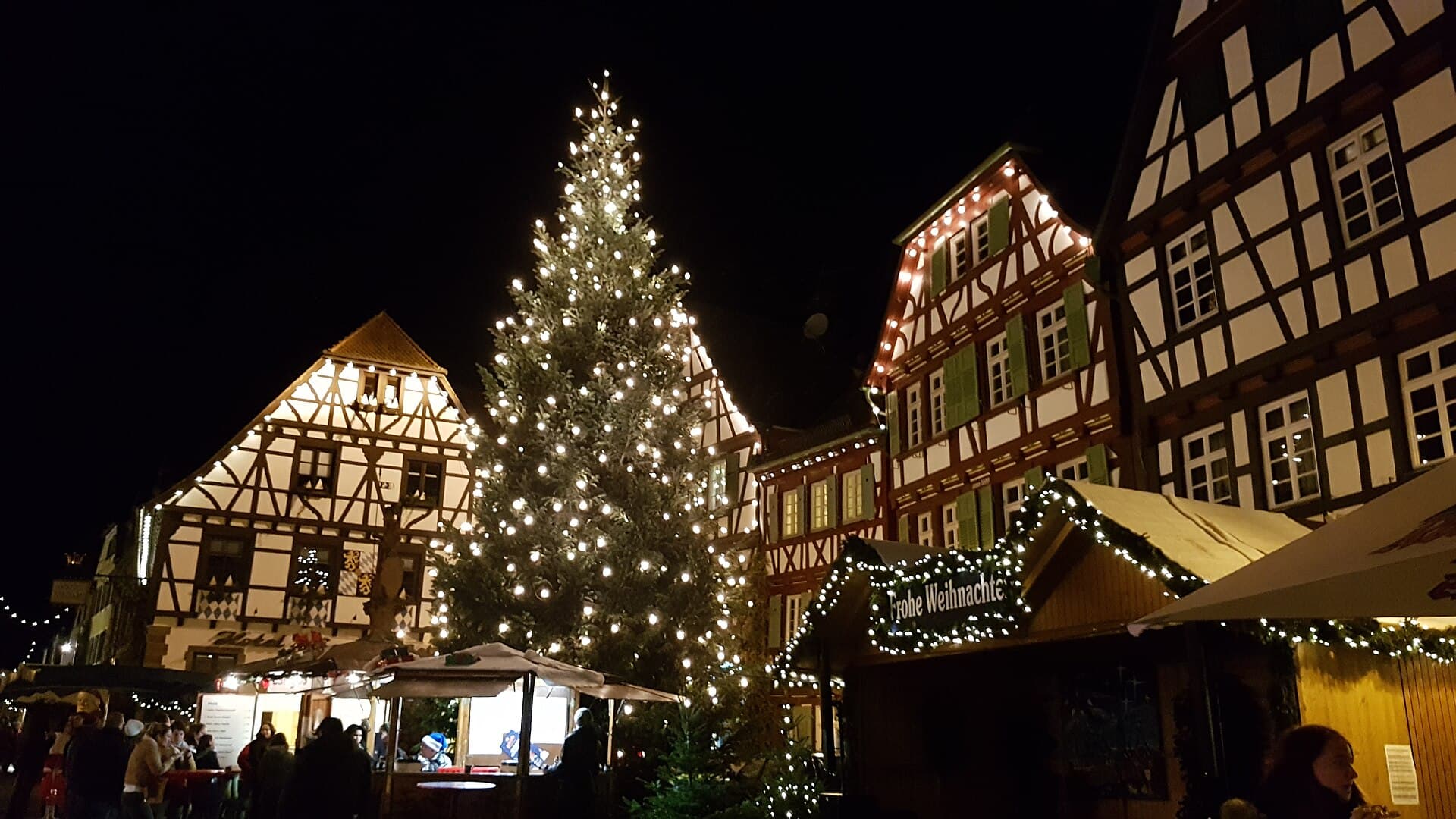 Weihnachtsmarkt auf dem Marktplatz in Bretten an einem Dezemberabend: ein beleuchteter Christbaum vor Fachwerkhäusern mit grünen Fensterläden, davor Holzhütten mit Lichterketten und Besucher an den Ständen.