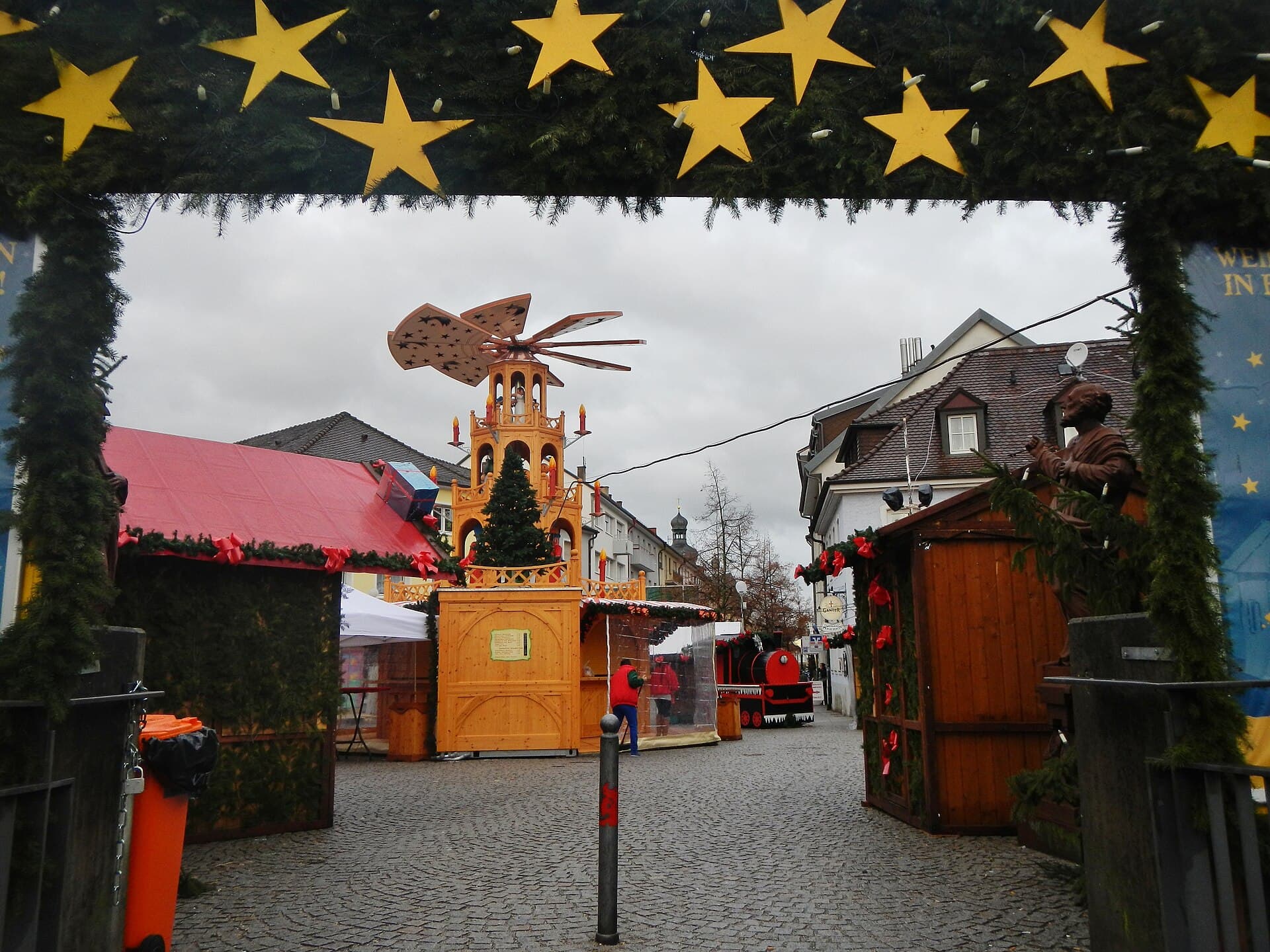 Blick durch einen Torbogen aus Tannengrün mit gelben Holzsternen auf den Weihnachtsmarkt am Otto-Oppenheimer-Platz in Bruchsal an einem Dezembervormittag, mit großer Weihnachtspyramide, Holzhütten und einer roten Lok für Kinder.