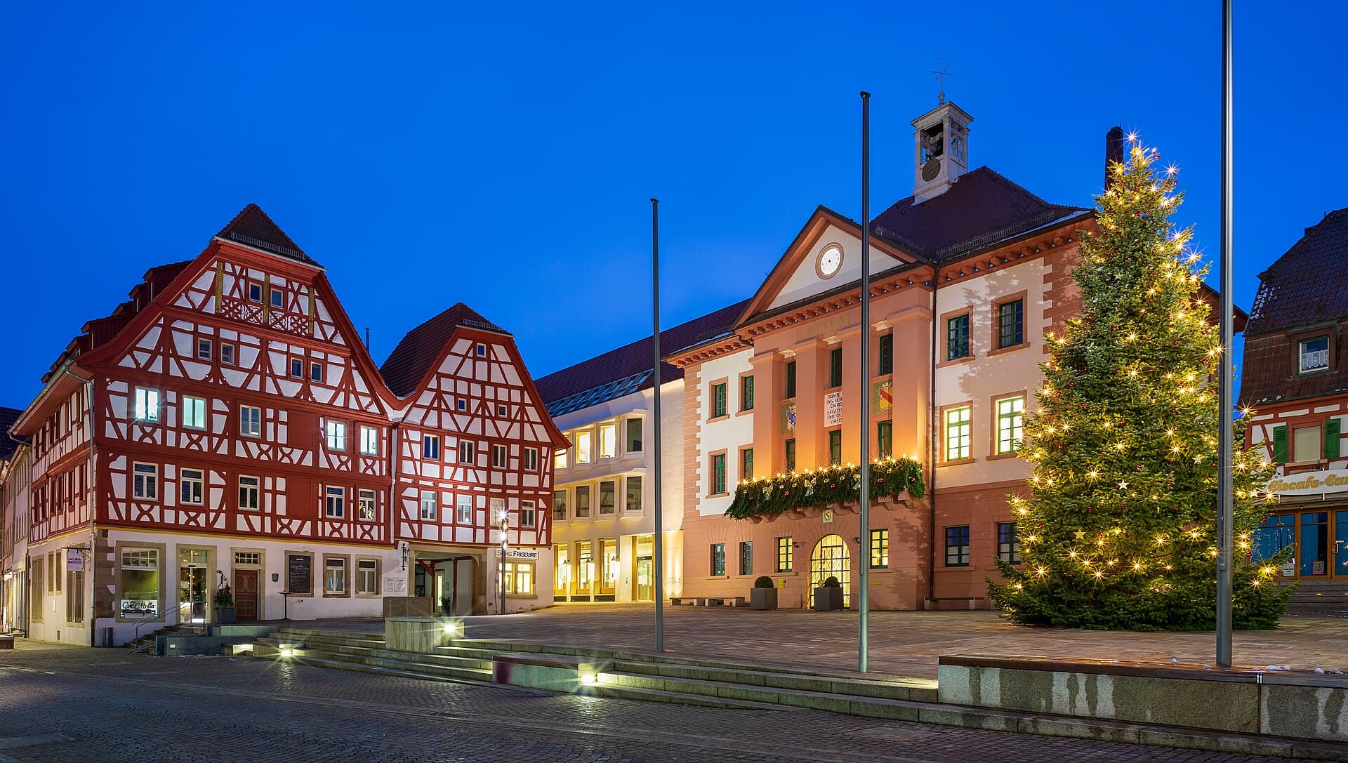 Der Marktplatz von Eppingen an einem Winterabend zur blauen Stunde: links zwei rot-weiße Fachwerkhäuser, in der Mitte ein Teil des Neuen und das Alte Rathaus mit Glockentürmchen und Lichtergirlande, rechts ein beleuchteter Christbaum.