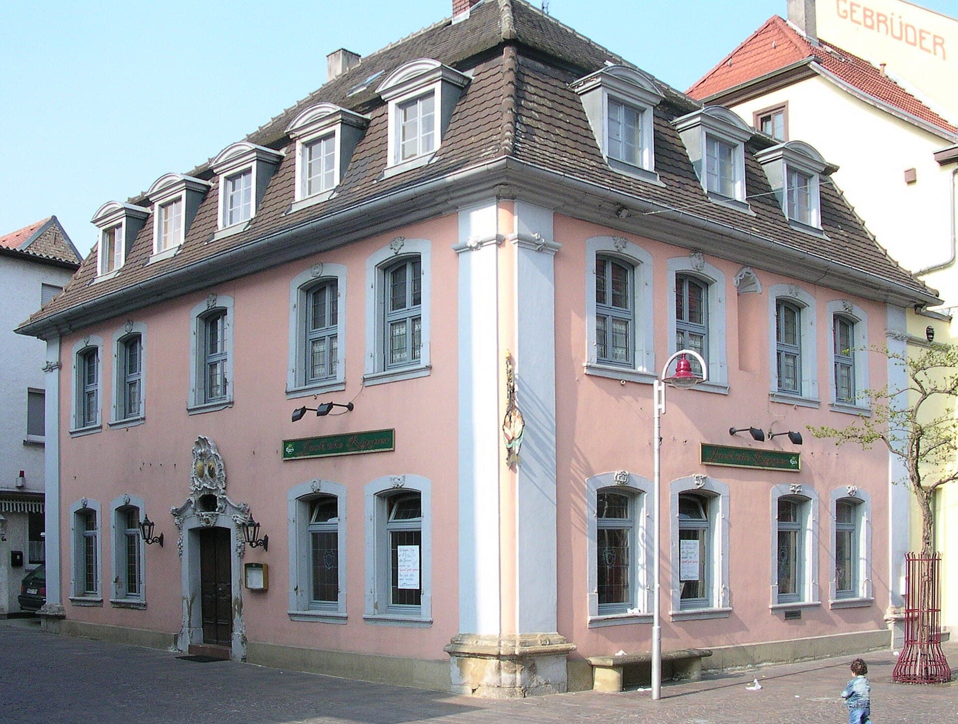 Das ehemalige katholische Pfarrhaus an der Ecke von Bahnhofstraße und Kirchplatz in Sinsheim, ein rosa verputzter Barockbau mit grauen Fensterrahmen, Mansarddach mit Gauben und verziertem Portal, davor das Pflaster der Fußgängerzone.