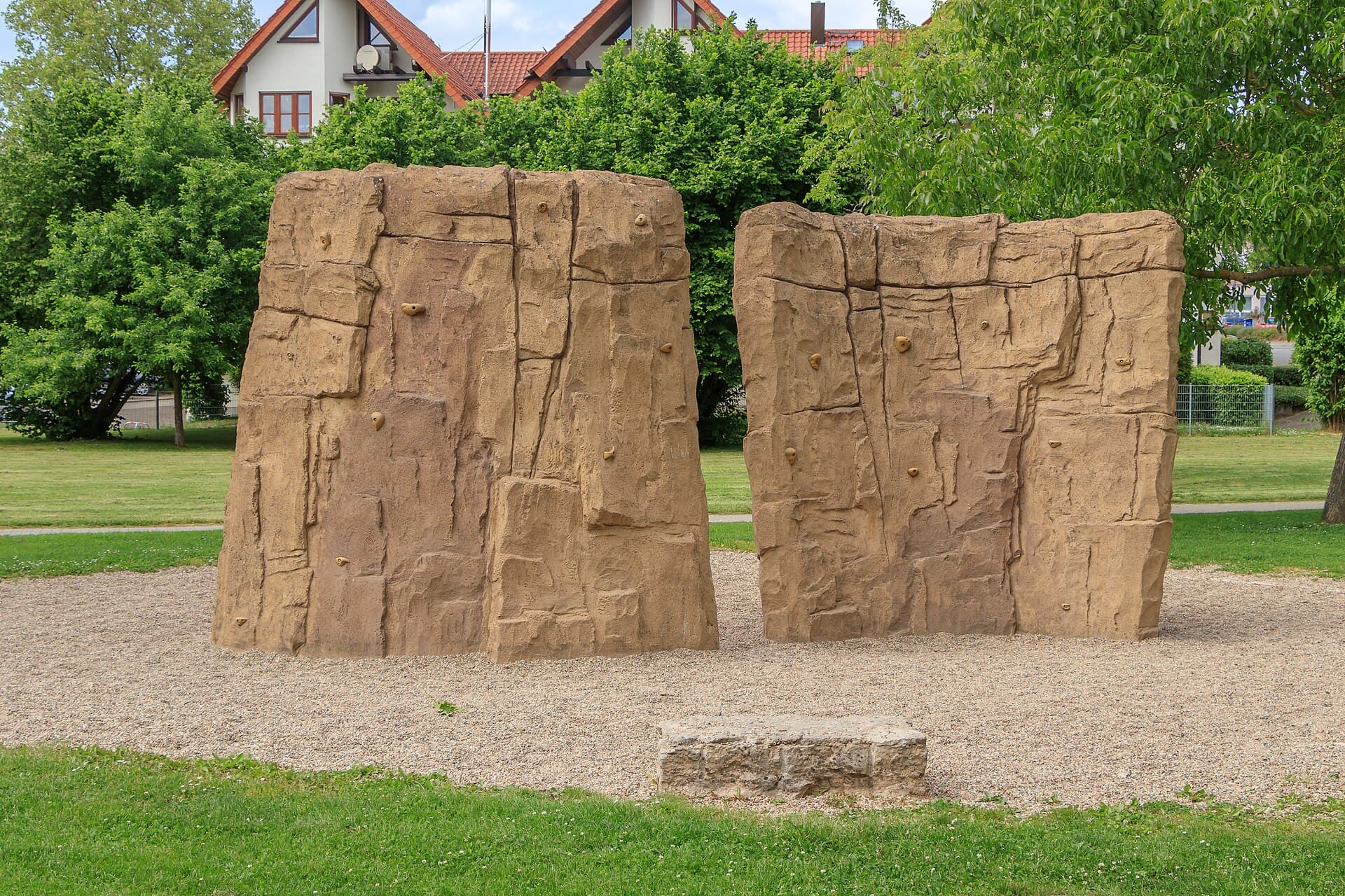 Zwei Kletterfelsen aus Kunststein auf einem Kiesbett im öffentlichen Spielplatz am Mühlbach in Bad Rappenau, dahinter Wiese, Bäume und Wohnhäuser unter blauem Himmel
