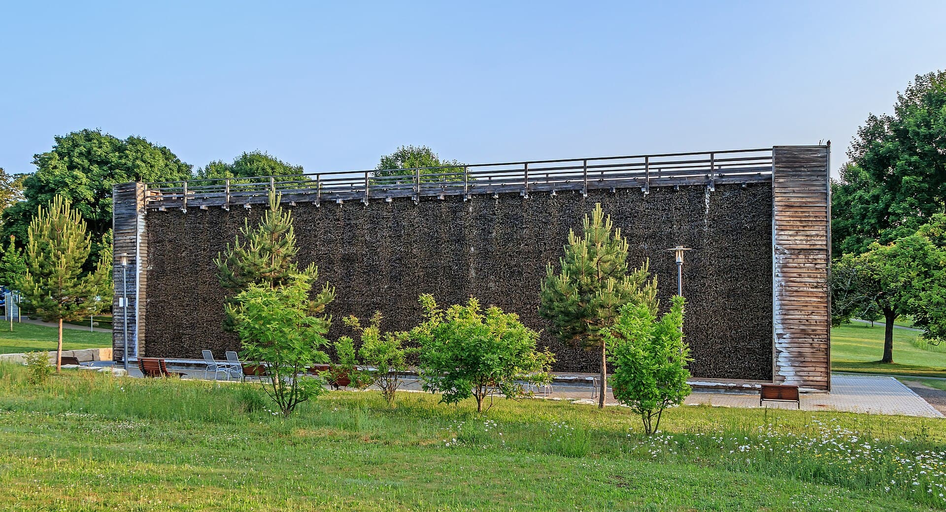 Das Gradierwerk im Salinenpark Bad Rappenau im Morgenlicht: eine lange dunkle Wand aus Schlehenreisig mit hölzernen Ecktürmen, davor junge Bäume und eine Wiese
